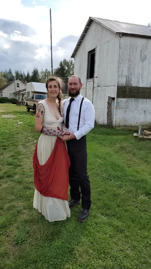 Stephanie in her wedding dress with her husband. Dress is comprised of an ecru linen bodice, with ecru silk chiffon skirt and red silk chiffon overdrape. A wool and silk band joins the bodice to the dress. Styled with wool arm warmers covered in white leaf pattern.