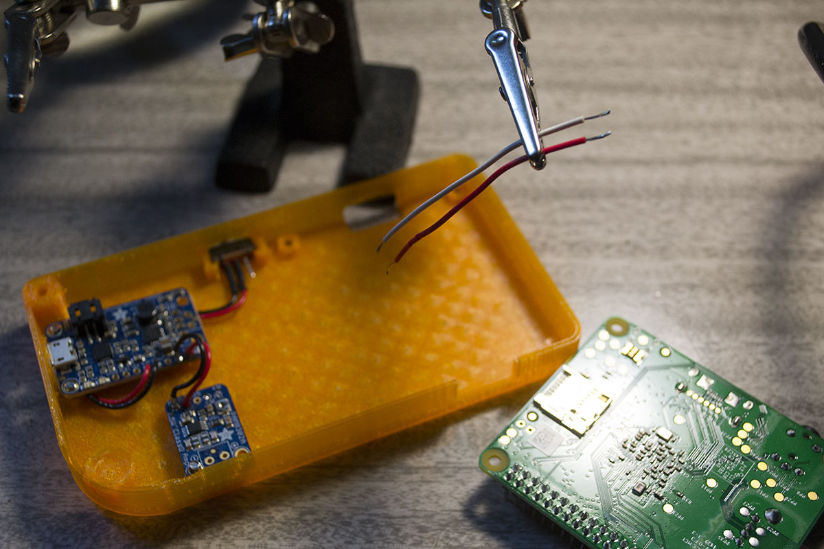 the pi grrl in progress, an orange half of the case contains the audio and power boost boards, and the raspberry pie lies beside it. A helping hand alligator clip holds two small wires in the foreground.